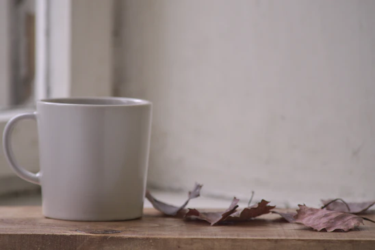 A close-up of a bright, hand-thrown ceramic mug resting beside autumn leaves on a wooden table.