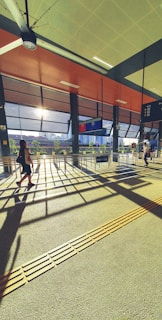 A sunlit transportation terminal with modern architectural design, featuring large windows that let in natural light. Two people walk through the space, casting long shadows on the textured floor. Information boards and signage are visible, suggesting a bus or train station.