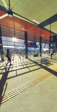 A sunlit transportation terminal with modern architectural design, featuring large windows that let in natural light. Two people walk through the space, casting long shadows on the textured floor. Information boards and signage are visible, suggesting a bus or train station.
