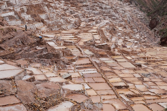 A sprawling landscape of terraced salt pans cascading down a hillside. The rectangular pools vary in color from light beige to deeper brown, indicating different stages of salt crystallization. The terrain is rugged with a complex network of pathways between the pools, and a few individuals are visible, engaged in the maintenance or harvesting of the salt. The surrounding area is earthy and mountainous.