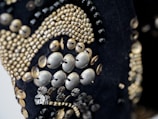 A close-up of intricate beadwork, featuring a variety of small, metallic beads and buttons arranged in decorative patterns. The beads are predominantly gold and silver, sewn onto a dark fabric background. The design appears ornate and elaborate, with different sizes and shapes creating texture and depth.