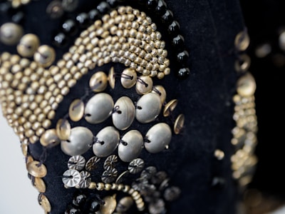 A close-up of intricate beadwork, featuring a variety of small, metallic beads and buttons arranged in decorative patterns. The beads are predominantly gold and silver, sewn onto a dark fabric background. The design appears ornate and elaborate, with different sizes and shapes creating texture and depth.