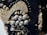 A close-up of intricate beadwork, featuring a variety of small, metallic beads and buttons arranged in decorative patterns. The beads are predominantly gold and silver, sewn onto a dark fabric background. The design appears ornate and elaborate, with different sizes and shapes creating texture and depth.