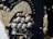 A close-up of intricate beadwork, featuring a variety of small, metallic beads and buttons arranged in decorative patterns. The beads are predominantly gold and silver, sewn onto a dark fabric background. The design appears ornate and elaborate, with different sizes and shapes creating texture and depth.