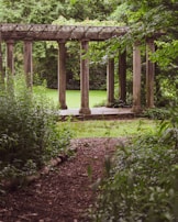 A serene garden scene with a classical stone pergola surrounded by lush greenery. The pathway leading towards the structure is bordered by dense vegetation, and the grass in the background is vibrant and well-maintained.