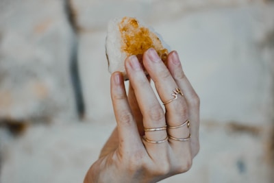 A hand adorned with multiple delicate rings is holding a rough, amber-colored crystal against a blurred, neutral background.