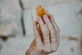 A hand adorned with multiple delicate rings is holding a rough, amber-colored crystal against a blurred, neutral background.