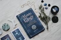 A dark blue book titled 'Moon Magic' lies on a textured fabric surface, accompanied by various mystical items such as gemstones, a small branch of herbs, and a pendant necklace. There are also cards with mystical imagery and a decorative stone nearby, conveying a sense of the occult and spirituality.