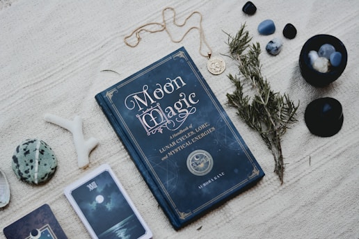 A dark blue book titled 'Moon Magic' lies on a textured fabric surface, accompanied by various mystical items such as gemstones, a small branch of herbs, and a pendant necklace. There are also cards with mystical imagery and a decorative stone nearby, conveying a sense of the occult and spirituality.