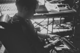 A black and white photograph of a person working in a music studio. The individual is seated, using a music production device with a mixing console and synthesizers visible in the background. The lighting is low, creating a moody atmosphere.