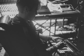 A black and white photograph of a person working in a music studio. The individual is seated, using a music production device with a mixing console and synthesizers visible in the background. The lighting is low, creating a moody atmosphere.
