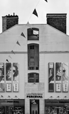 A black and white image of a building facade with two large posters displaying people wearing glasses. The building has the words 'Les Opticiens Perceval' above the entrance. Triangular flags are strung from above, crossing in front of the building.
