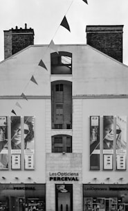 A black and white image of a building facade with two large posters displaying people wearing glasses. The building has the words 'Les Opticiens Perceval' above the entrance. Triangular flags are strung from above, crossing in front of the building.