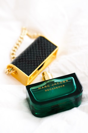A green bottle of Marc Jacobs Decadence perfume sits on a light surface, with a golden and black-textured accessory in the background. The perfume bottle is elegantly designed with a rectangular shape and golden cap.