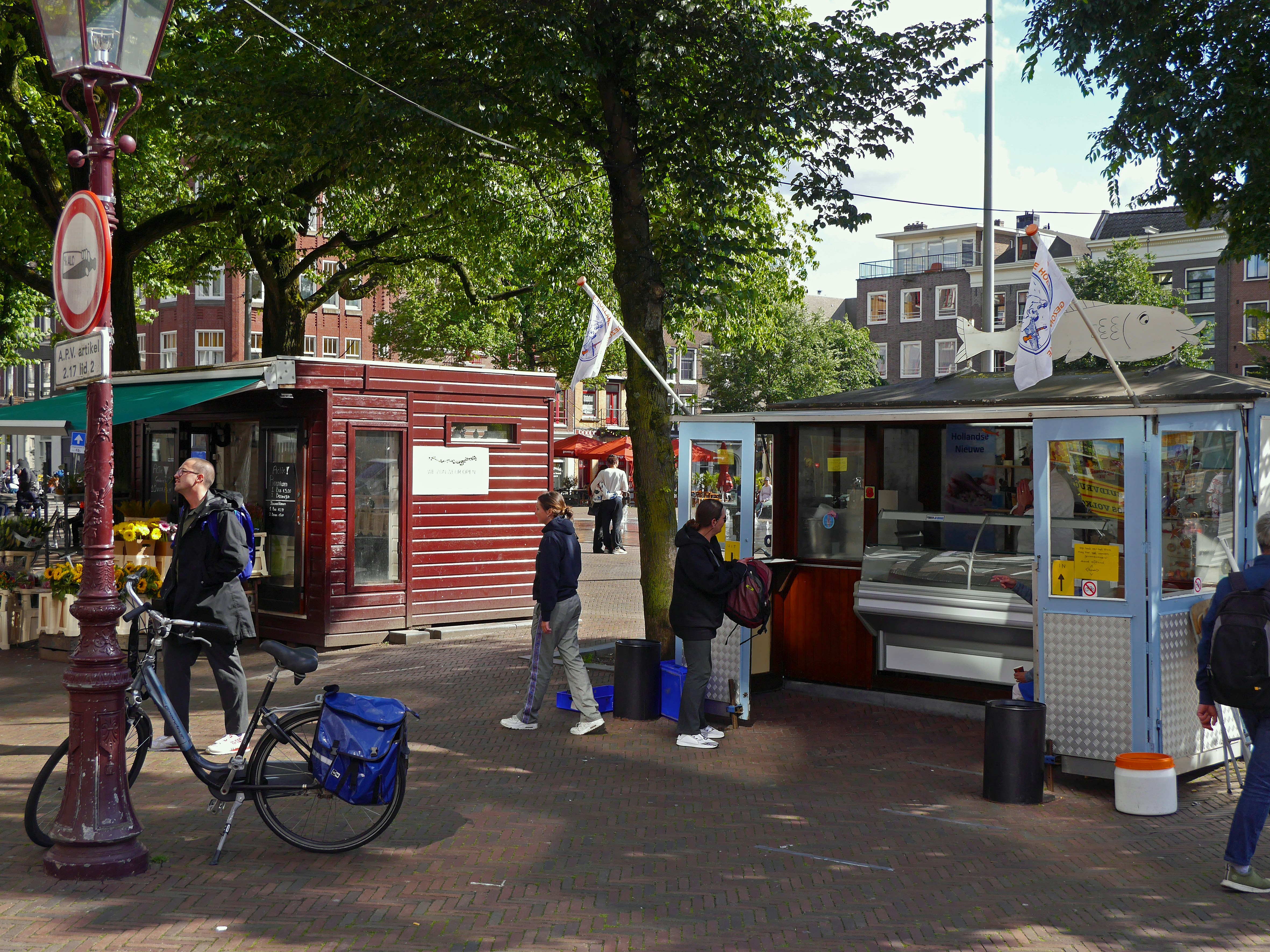 Cozy square in Amsterdam old city with people eating and walking in the street. There is a fish stall and a flower stable, and for children a fountain with water jets,coming from out the brick pavement. Free photo of Dutch street view of 10 June 2020. Viskraam op het Haarlemmerplein en bloemenstal; gratis foto Amsterdam, Fons Heijnsbroek