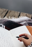 A hand holding a black pen is poised to write in an open notebook filled with cursive writing. The notebook rests on a surface covered with a patterned blanket, suggesting a cozy environment. The background features a wooden texture, possibly the edge of a porch or a rustic table.