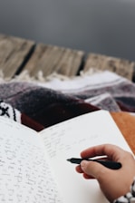 A hand holding a black pen is poised to write in an open notebook filled with cursive writing. The notebook rests on a surface covered with a patterned blanket, suggesting a cozy environment. The background features a wooden texture, possibly the edge of a porch or a rustic table.