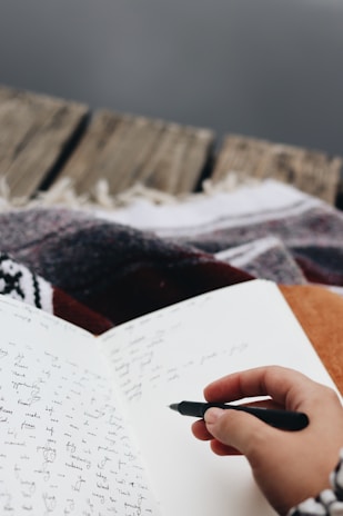 A hand holding a black pen is poised to write in an open notebook filled with cursive writing. The notebook rests on a surface covered with a patterned blanket, suggesting a cozy environment. The background features a wooden texture, possibly the edge of a porch or a rustic table.