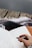 A hand holding a black pen is poised to write in an open notebook filled with cursive writing. The notebook rests on a surface covered with a patterned blanket, suggesting a cozy environment. The background features a wooden texture, possibly the edge of a porch or a rustic table.