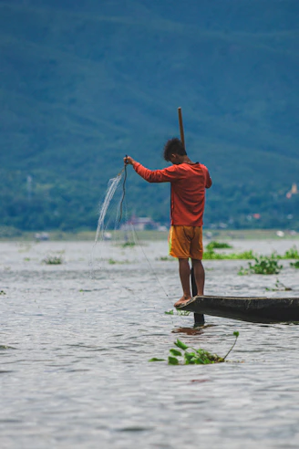 man in orange jacket and brown shorts standing on brown wooden log on body of water