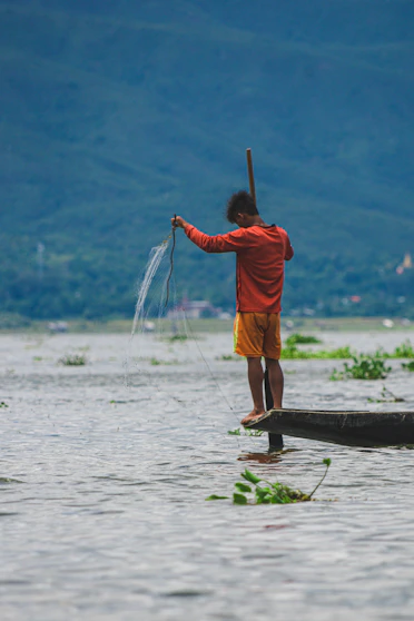 man in orange jacket and brown shorts standing on brown wooden log on body of water