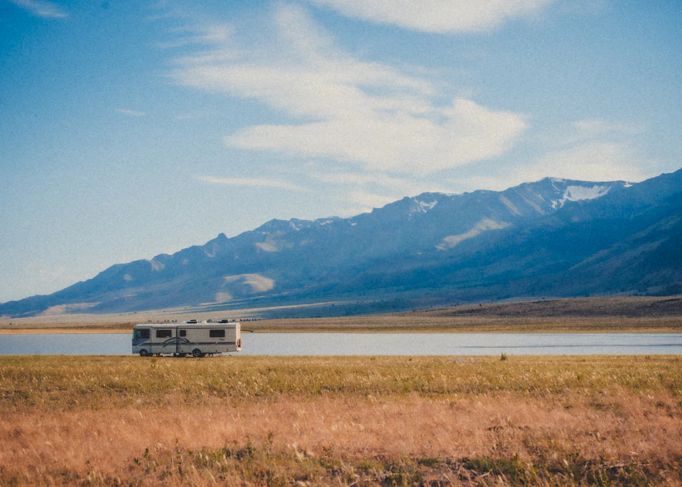 White motor home parked on lake shore amidst tall grass and distant mountains