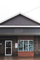 A single-story building with dark wooden siding and a large brick section with a window. The entrance door is on the left side with a sign indicating accessibility and a no-smoking sign. The window displays a neon 'OPEN' sign, and a reflection of nearby vehicles can be seen.