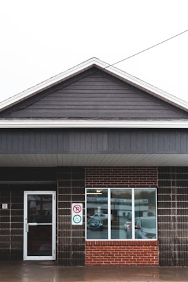 A single-story building with dark wooden siding and a large brick section with a window. The entrance door is on the left side with a sign indicating accessibility and a no-smoking sign. The window displays a neon 'OPEN' sign, and a reflection of nearby vehicles can be seen.