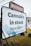 A large outdoor sign for Spiritleaf, a cannabis store, displays the message 'Cannabis in stock, Now Open' along with 'Locally Owned & Operated.' The sign is positioned on a grassy area with metal supports, and the background includes some buildings and power lines.