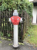 A fire hydrant with a silver metallic body and a red top, decorated with googly eyes, giving it a cartoonish appearance. It is set against a wooden fence with green foliage, on a paved pathway.
