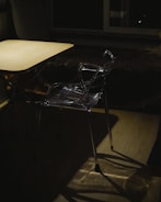 A clear plastic chair is positioned near a wooden table in a dimly lit room. Shadows are cast on the floor, and there is a large window in the background, adding an element of depth to the scene. The lighting creates a moody and somewhat mysterious atmosphere.