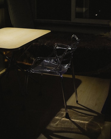 A clear plastic chair is positioned near a wooden table in a dimly lit room. Shadows are cast on the floor, and there is a large window in the background, adding an element of depth to the scene. The lighting creates a moody and somewhat mysterious atmosphere.