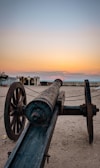 A vintage cannon rests on a sandy surface, facing towards the horizon where a warm sunset transitions from orange to pale blue. The scene includes an old building with columns and archways, situated in the background. The environment appears calm and historical.