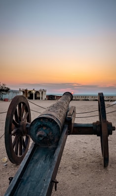 A vintage cannon rests on a sandy surface, facing towards the horizon where a warm sunset transitions from orange to pale blue. The scene includes an old building with columns and archways, situated in the background. The environment appears calm and historical.
