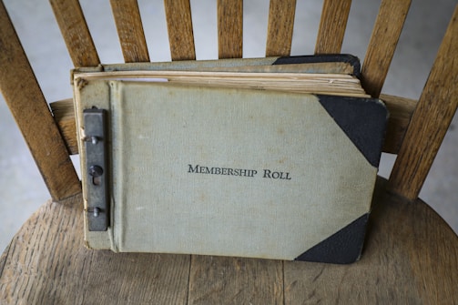 A vintage membership roll book lies on a wooden chair. The book has a worn, textured cover with the title written on the front. The binding appears to be metal, showing signs of age and use.