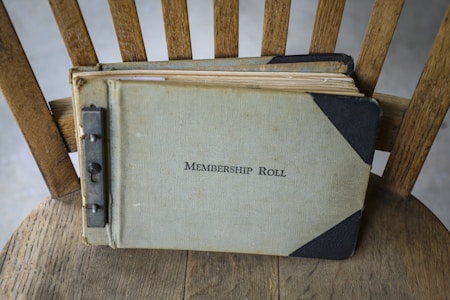 A vintage membership roll book lies on a wooden chair. The book has a worn, textured cover with the title written on the front. The binding appears to be metal, showing signs of age and use.
