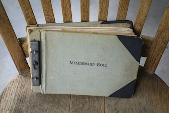 A vintage membership roll book lies on a wooden chair. The book has a worn, textured cover with the title written on the front. The binding appears to be metal, showing signs of age and use.