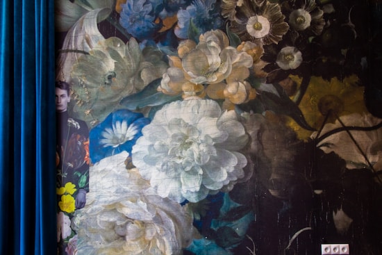 A richly detailed floral mural with a mix of large, lush flowers in varying shades, primarily whites, blues, and yellows, fills the main area. A man peeks from behind a blue velvet curtain on the left edge, partially in view.