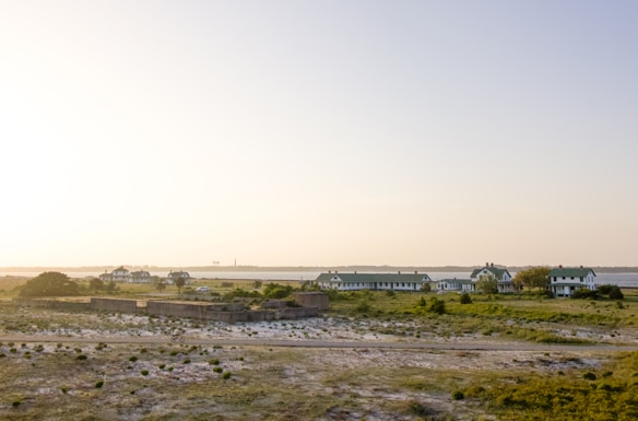 A serene landscape featuring a cluster of green-roofed buildings amidst a flat, grassy terrain. The sky is clear with a pale gradient, and there is a body of water in the background. Sparse vegetation is scattered across the sandy foreground, lending a sense of tranquility to the setting.