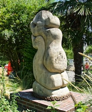 A large, abstract stone sculpture stands amidst lush greenery, including tall palm and bamboo plants. The sculpture is mounted on a brick platform with a metal chain encircling its base. Intricate carvings depicting a boat and waves are visible on the surface of the sculpture.