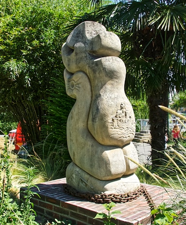A large, abstract stone sculpture stands amidst lush greenery, including tall palm and bamboo plants. The sculpture is mounted on a brick platform with a metal chain encircling its base. Intricate carvings depicting a boat and waves are visible on the surface of the sculpture.