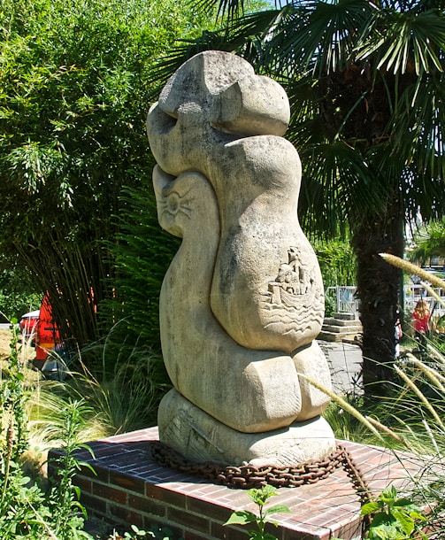 A large, abstract stone sculpture stands amidst lush greenery, including tall palm and bamboo plants. The sculpture is mounted on a brick platform with a metal chain encircling its base. Intricate carvings depicting a boat and waves are visible on the surface of the sculpture.