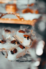 White chocolate covered treats garnished with whole almonds and small decorative sugar balls are presented on a reflective surface with intricate gold patterns. The setting suggests an elegant and festive presentation, with soft bokeh effects in the background adding to the luxurious feel.