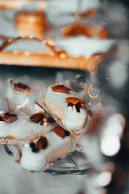 White chocolate covered treats garnished with whole almonds and small decorative sugar balls are presented on a reflective surface with intricate gold patterns. The setting suggests an elegant and festive presentation, with soft bokeh effects in the background adding to the luxurious feel.