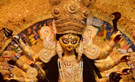 An intricately adorned statue of a Hindu goddess with multiple arms, each holding various symbolic items, surrounded by rich golden decorations and intricate detailing. The background includes traditional art scenes and vivid colors.