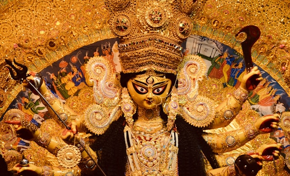 An intricately adorned statue of a Hindu goddess with multiple arms, each holding various symbolic items, surrounded by rich golden decorations and intricate detailing. The background includes traditional art scenes and vivid colors.
