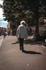 An elderly person with white hair is wearing a light blue jacket and walking down a paved street shaded by trees. The scene captures an urban environment with old buildings in the background and a few other people walking in the distance. The sunlight filtering through the tree leaves creates a complex pattern of light and shadow on the ground.