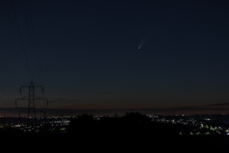 A night sky featuring a visible comet with a bright tail, above a cityscape with twinkling lights on the horizon. A silhouetted power line tower is visible on the left side, indicating a rural or suburban area.