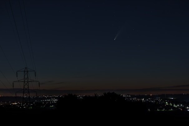 A night sky featuring a visible comet with a bright tail, above a cityscape with twinkling lights on the horizon. A silhouetted power line tower is visible on the left side, indicating a rural or suburban area.