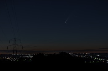 A night sky featuring a visible comet with a bright tail, above a cityscape with twinkling lights on the horizon. A silhouetted power line tower is visible on the left side, indicating a rural or suburban area.